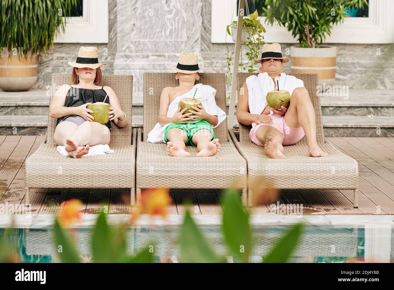 Father, mother and son covering faces with hats when relaxing on chaise-lounges with coconut cocktails in hands Stock Photo