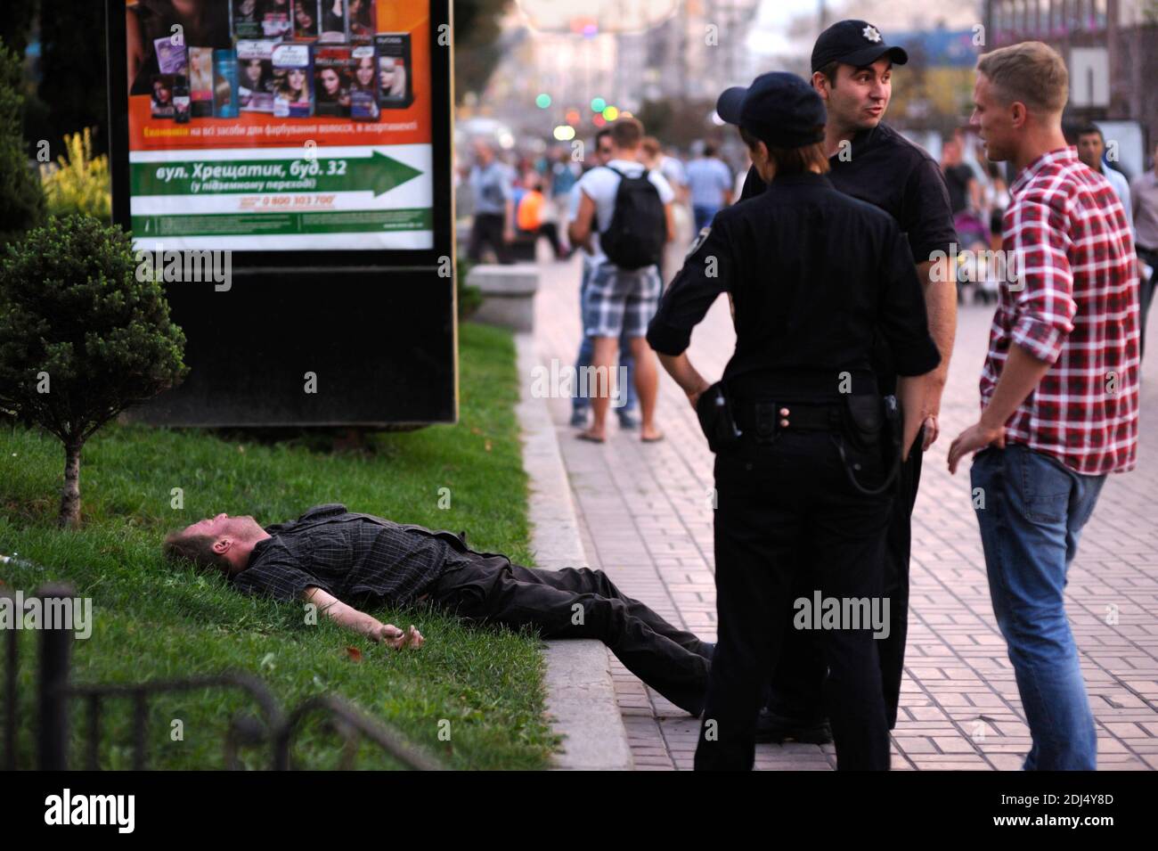 Very drunk man falling asleep on a street lawn, patrol of police talking to the young man Stock Photo