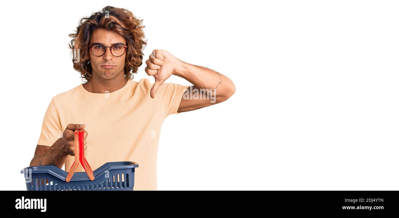 Young hispanic man holding supermarket shopping basket with angry face, negative sign showing dislike with thumbs down, rejection concept Stock Photo