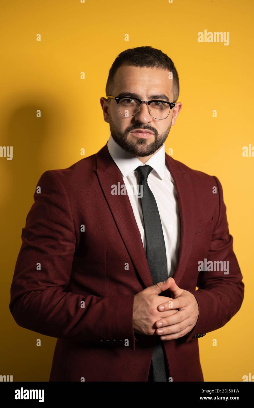 Half-length portrait of handsome man standing half turn in burgundy color suit and eye glasses looking at camera with hands folded isolated on yellow Stock Photo