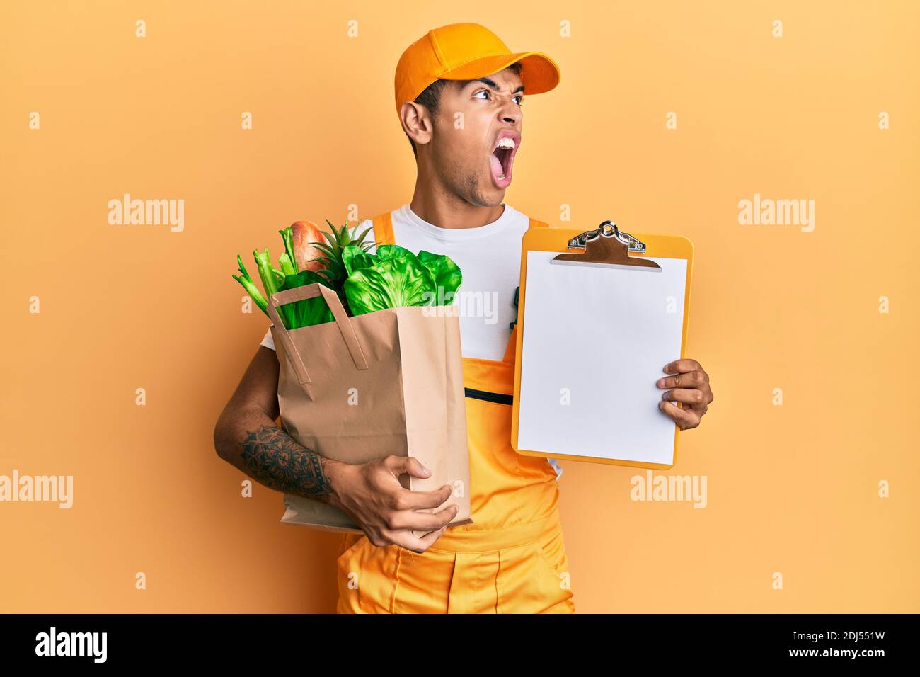 Young handsome african american man wearing courier uniform with groceries from supermarket and clipboard angry and mad screaming frustrated and furio Stock Photo