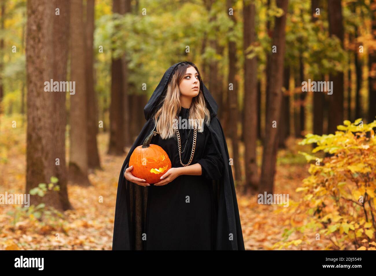 Young woman in black cape carrying spooky jack o lantern and looking away on Halloween day in autumn forest Stock Photo