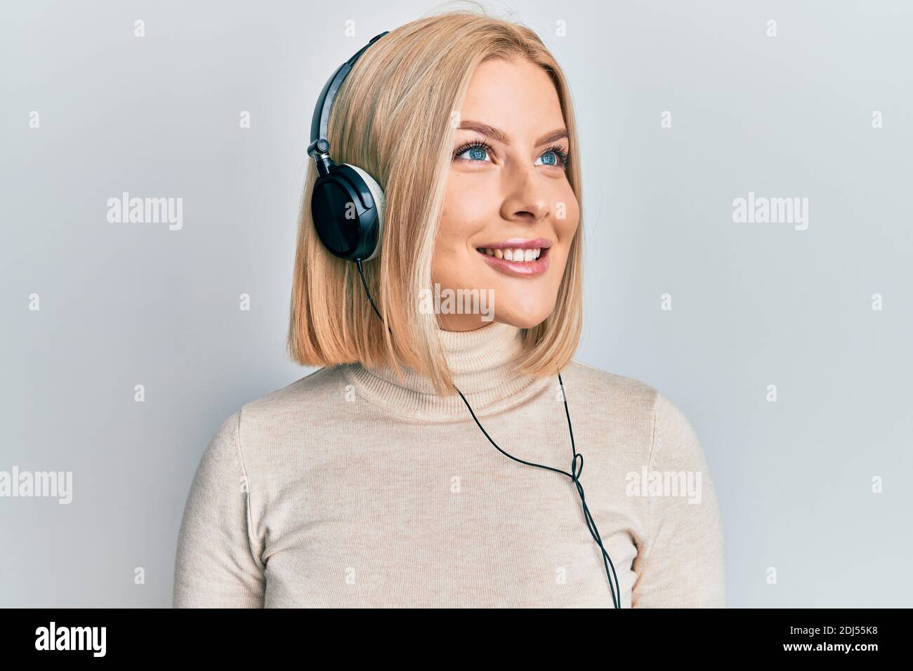 Young blonde woman listening to music using headphones looking away to ...