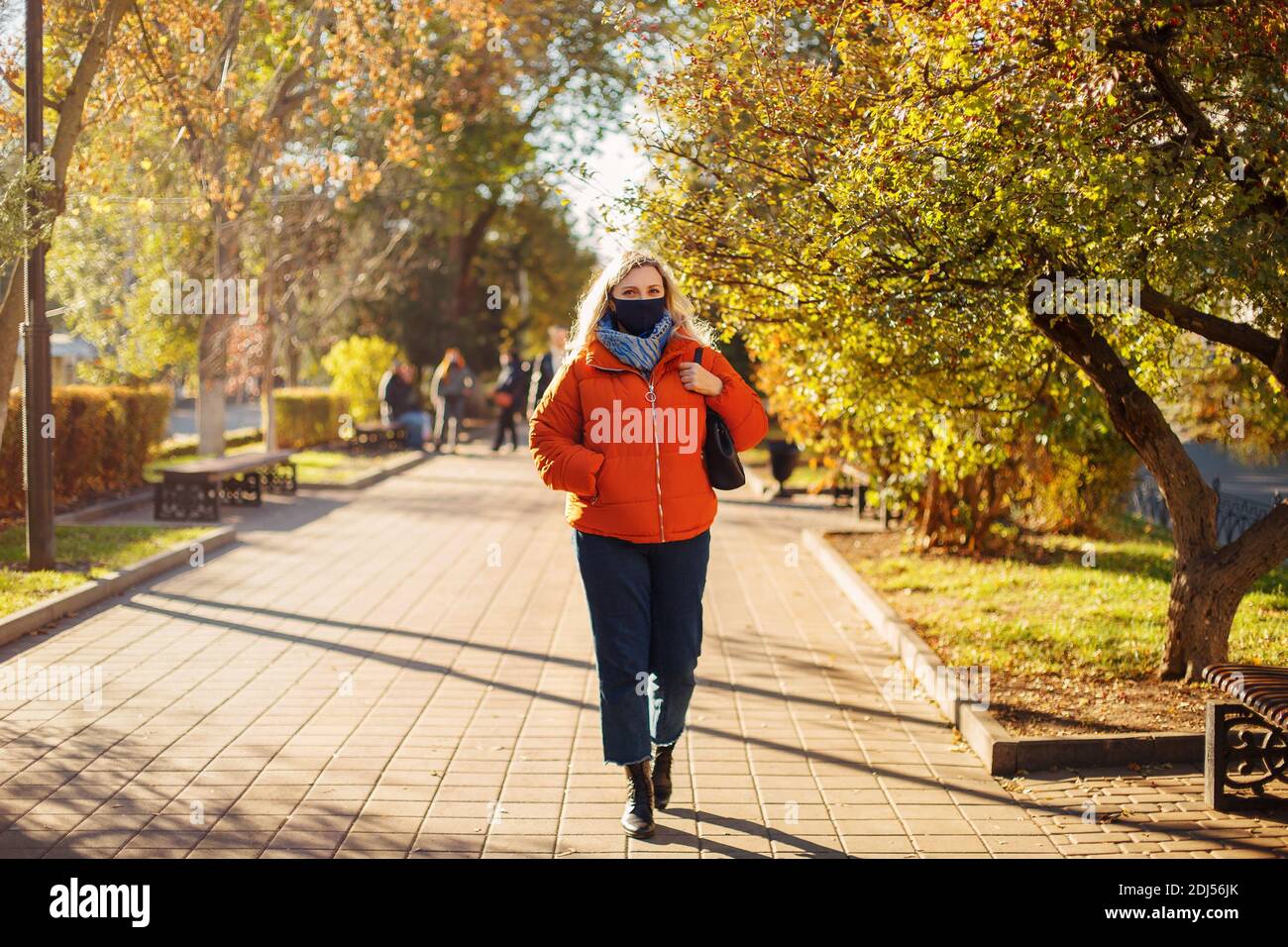 Full body woman in outerwear and mask walking on paved path on sunny day in autumn park during pandemic Stock Photo