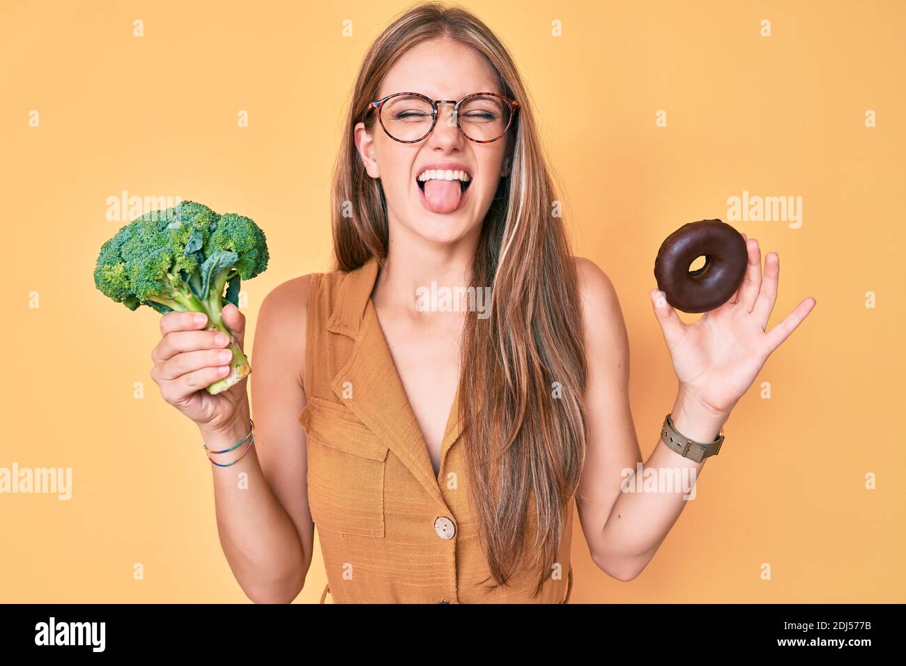 Young blonde girl holding broccoli and chocolate donut sticking tongue out happy with funny expression. Stock Photo