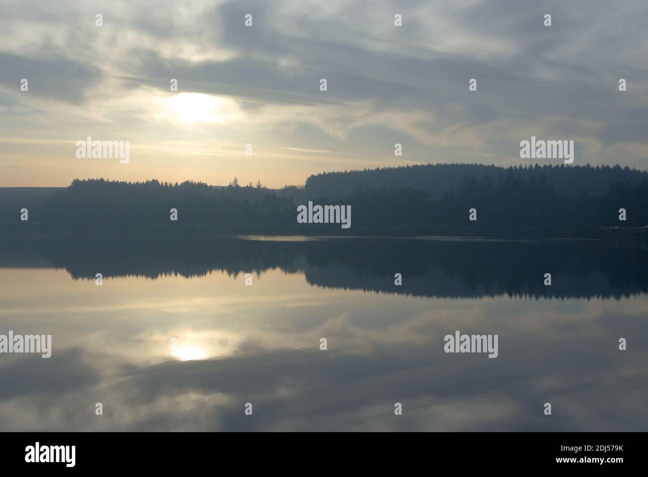 Winters sun and a cloudy sky reflected perfectly in the calm water of redmires reservoir. Hazy forest tree line in the distance, cars parked beneath Stock Photo