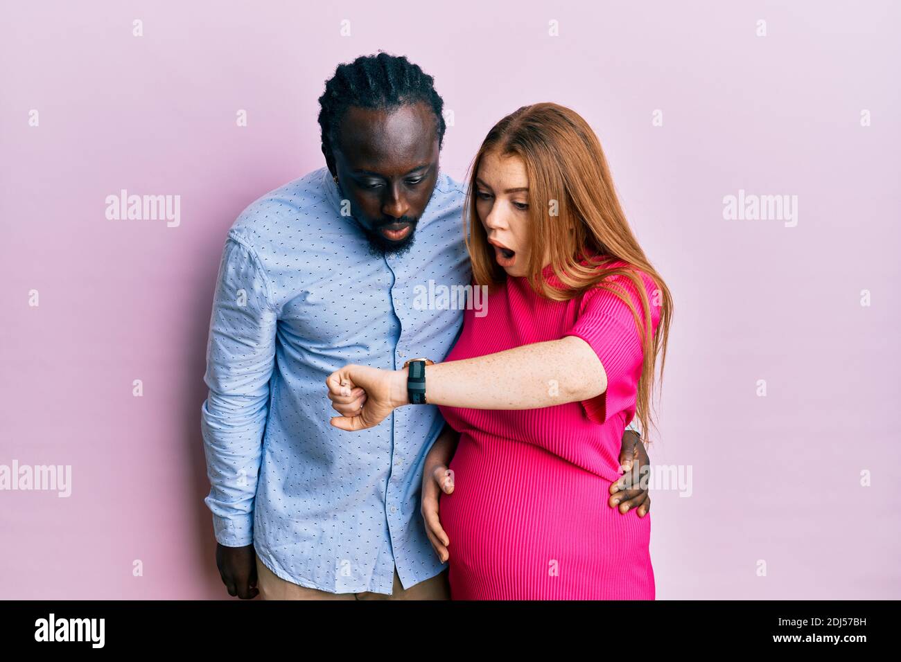 Young interracial couple expecting a baby, touching pregnant belly looking at the watch time worried, afraid of getting late Stock Photo