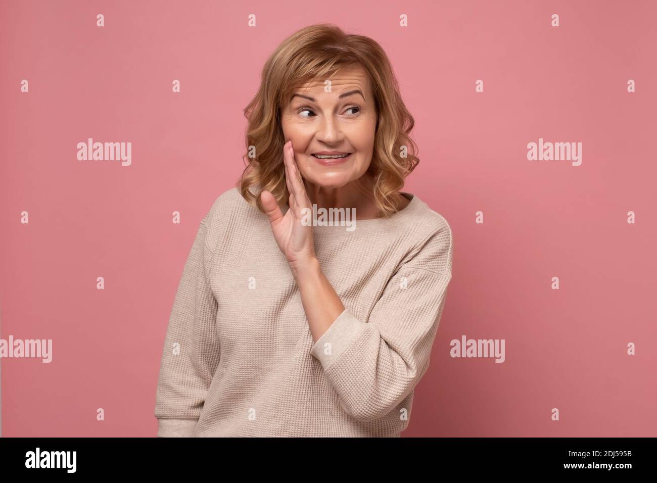 Senior woman with hand on mouth telling secret rumor, whispering malicious talk conversation. studio shot on pink wall. Stock Photo
