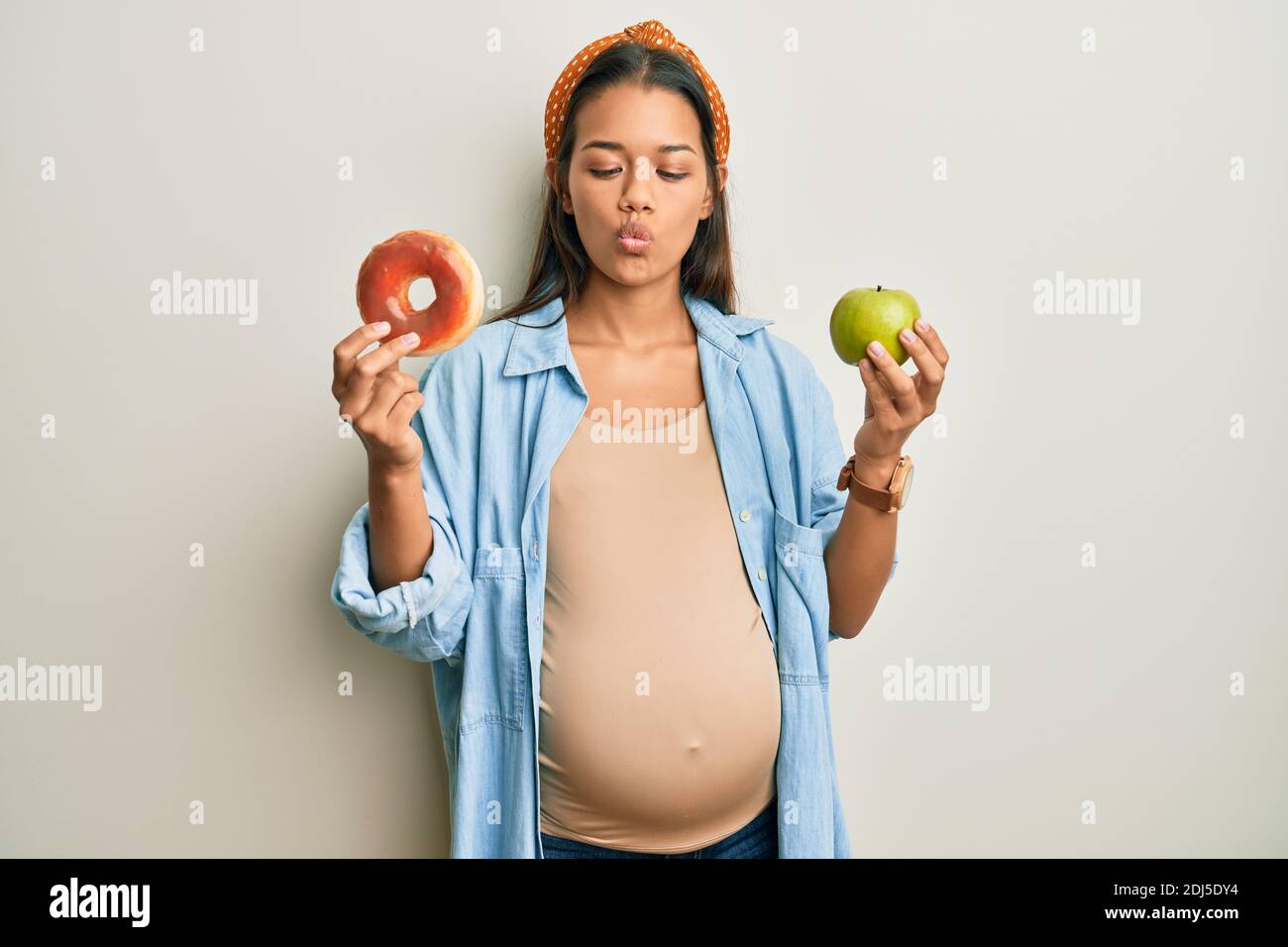 Beautiful hispanic woman expecting a baby, holding food making fish face with mouth and squinting eyes, crazy and comical. Stock Photo