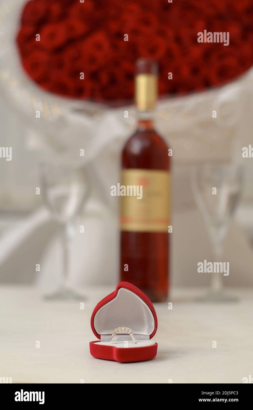 Silver ring in a red box in the shape of a heart on a white table on the background of a bottle of wine, empty glasses and a large bouquet of red rose Stock Photo