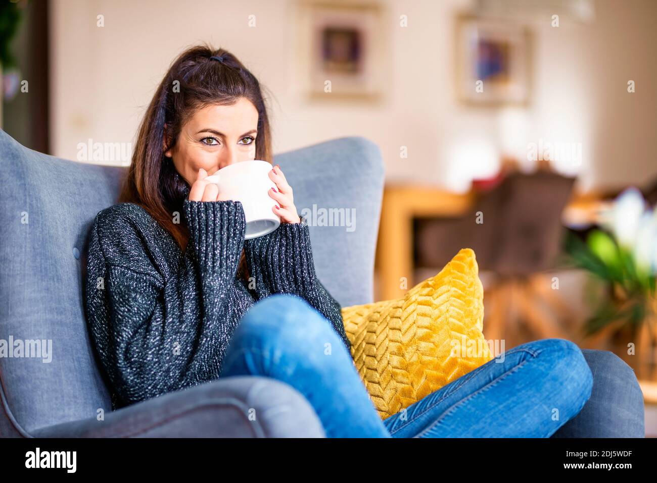 Portrait shot of smiling woman drinking tea while relaxing in the armchair at home. Stock Photo