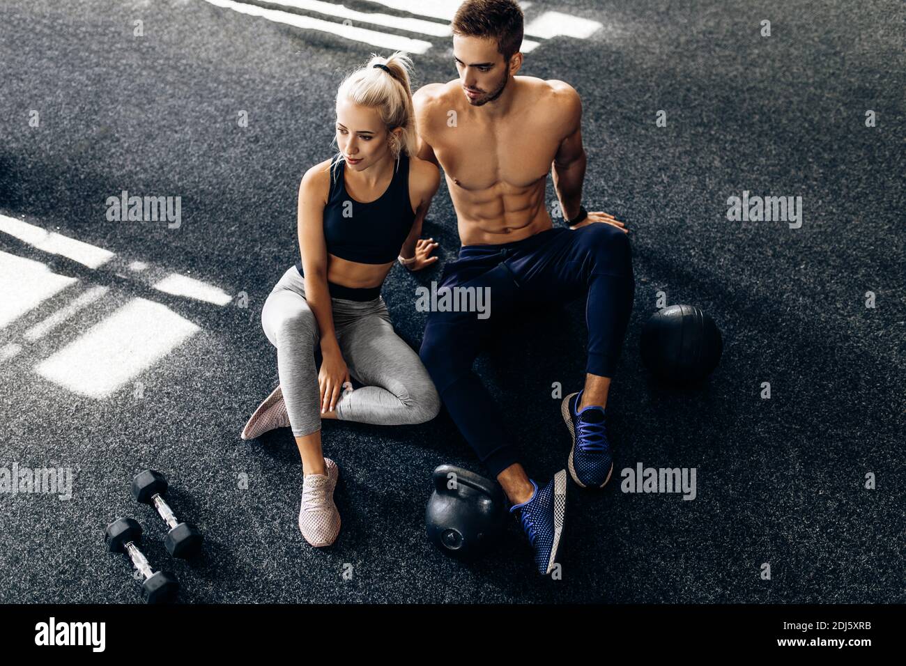 Young couple of athletic people, in sportswear, sitting together on the floor of the gym, with dumbbells, together after training in the gym Stock Photo
