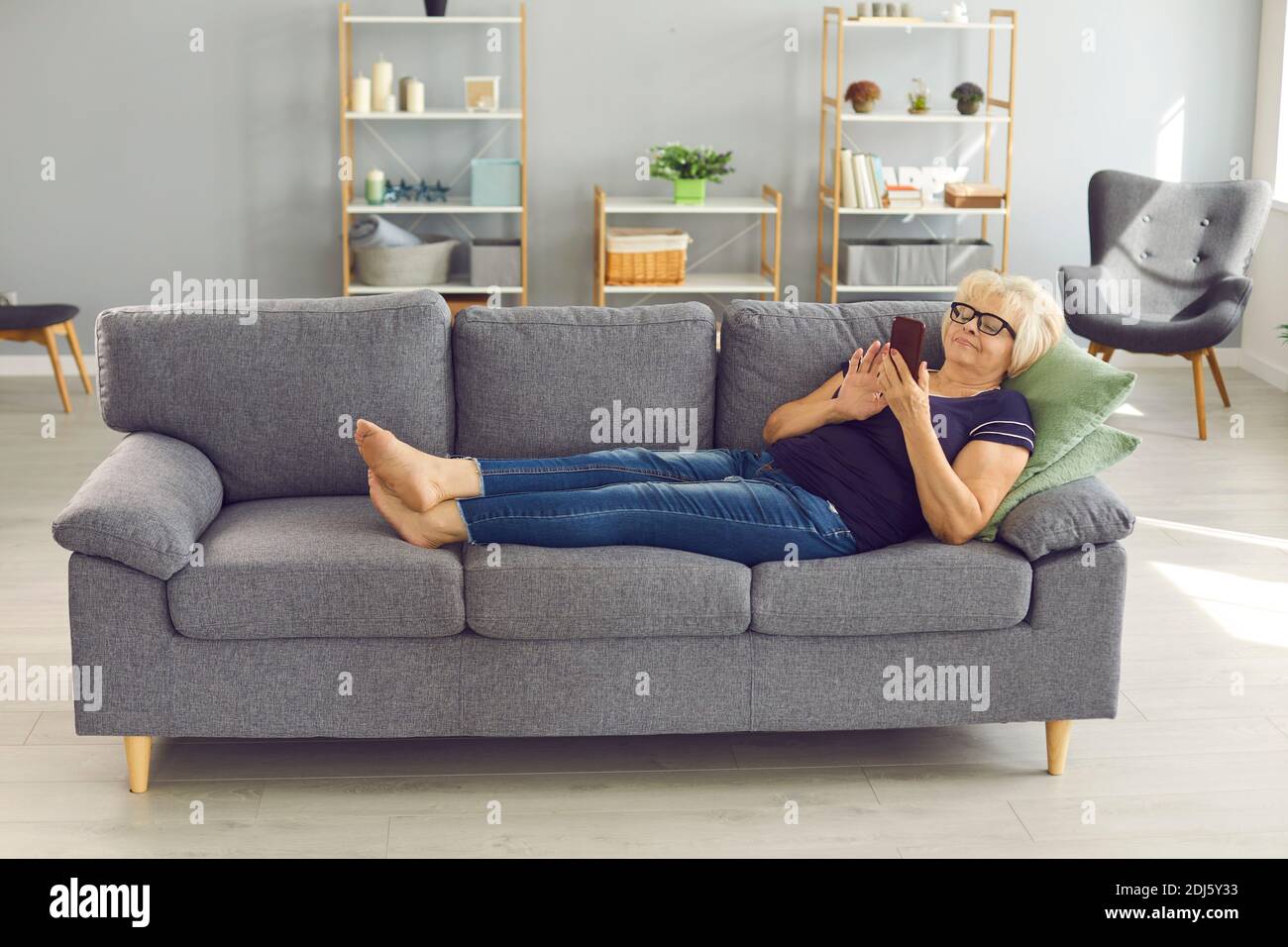 Positive senior woman lying on sofa, relaxing and chatting or making order online on smartphone Stock Photo