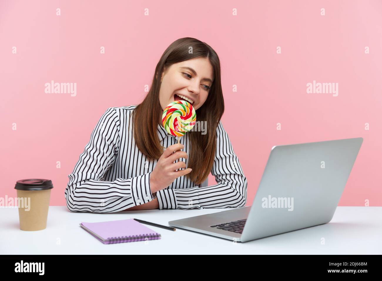 Funny playful woman licking and biting round colored lollipop on stick talking on video call at laptop sitting at workplace, having fun. Indoor studio Stock Photo