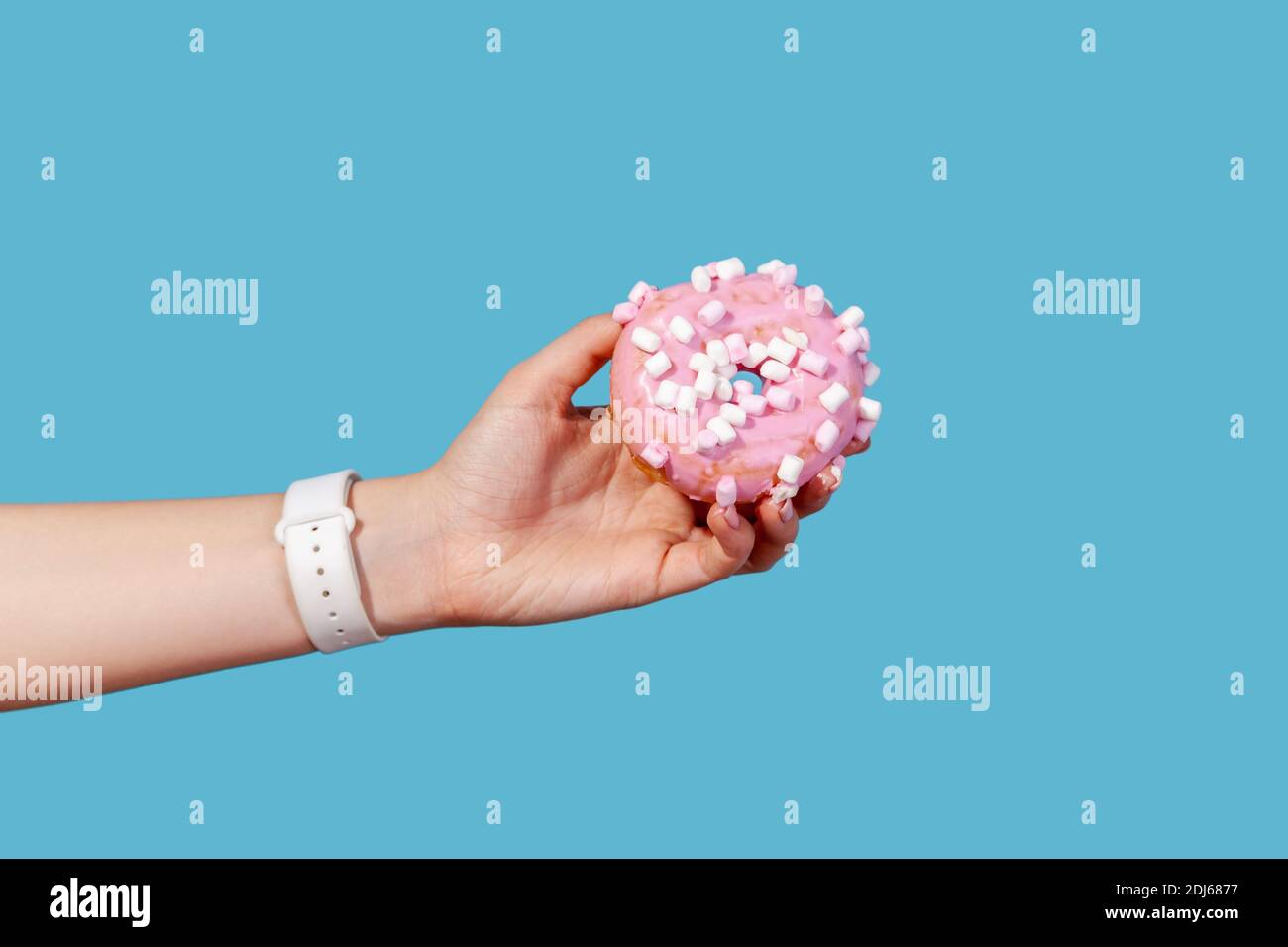Close up side view female hand holding sweet round donut with pink icing, delicious junk dessert saturated with fats and carbohydrates. Indoor studio Stock Photo