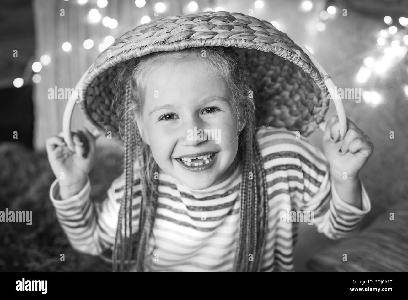 A mischievous laughing girl with pigtails and a wicker basket on her head against the background of a glowing garland. Stock Photo