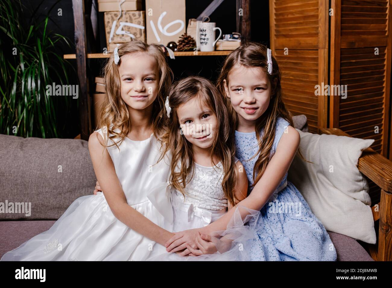 Two cute sisters in white dresses sitting on sofa close to each other and playing at home. Little sister hugging her smallest sister. Stock Photo