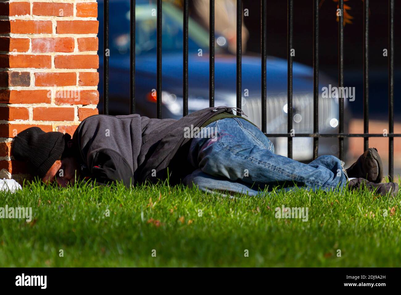Alexandria, VA, USA  11-28-2020: A homeless latino man wearing sweatshirt and jeans is sleeping on the grass outdoors at a city park by a brick wall a Stock Photo