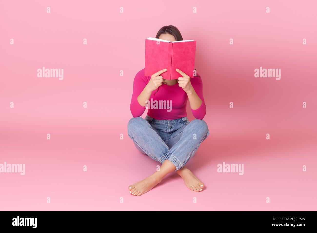 portrait of a young woman sitting cross-legged on the floor and hiding her face behind an open book and reading a book in her hands Stock Photo