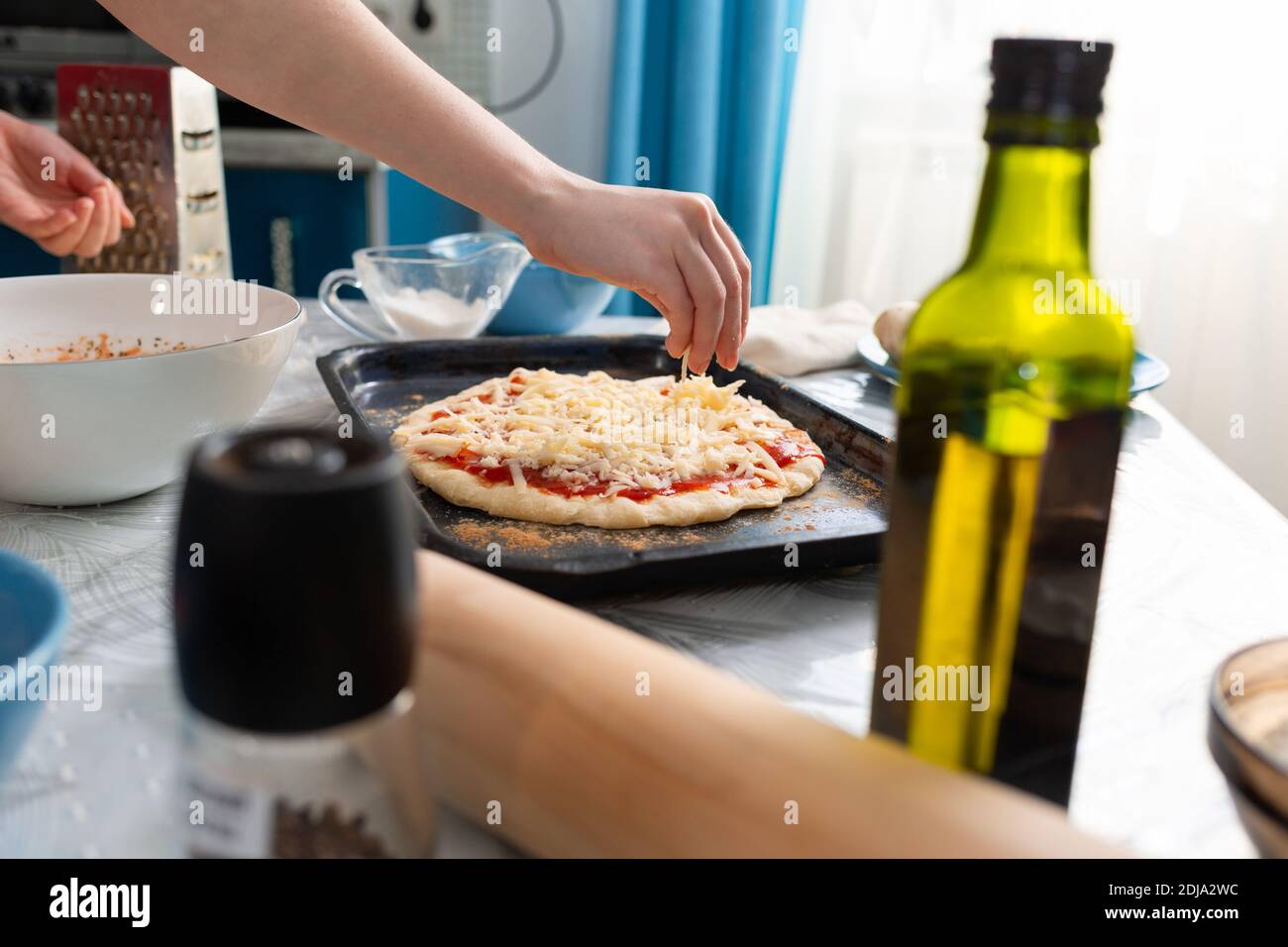A female's hand sprinkles cheese on a pizza tray. Close-up. Side view. Home cooking. Stock Photo