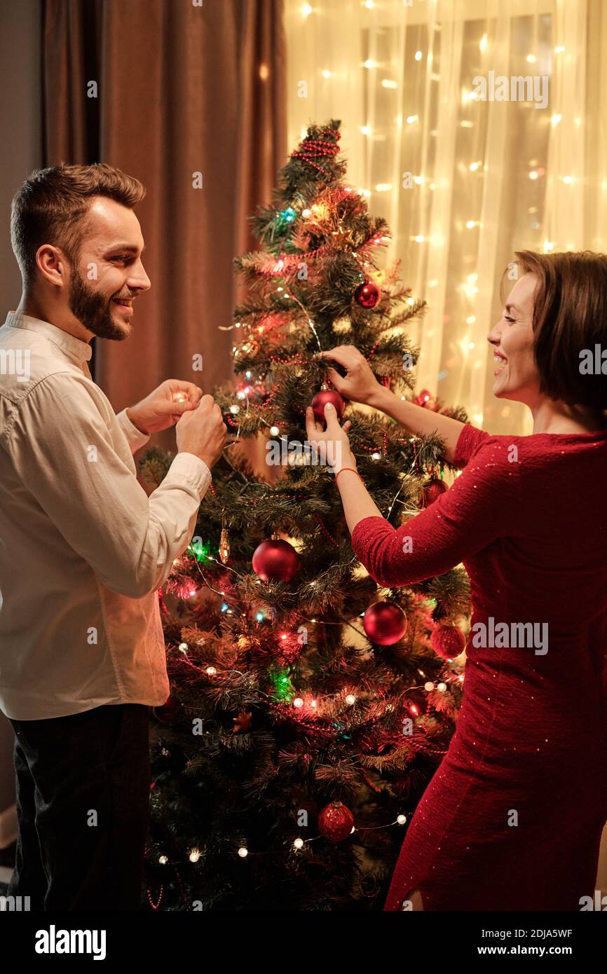 Rear view of cheerful beautiful young couple enjoying Christmas tree decorating and laughing while chatting in living room Stock Photo