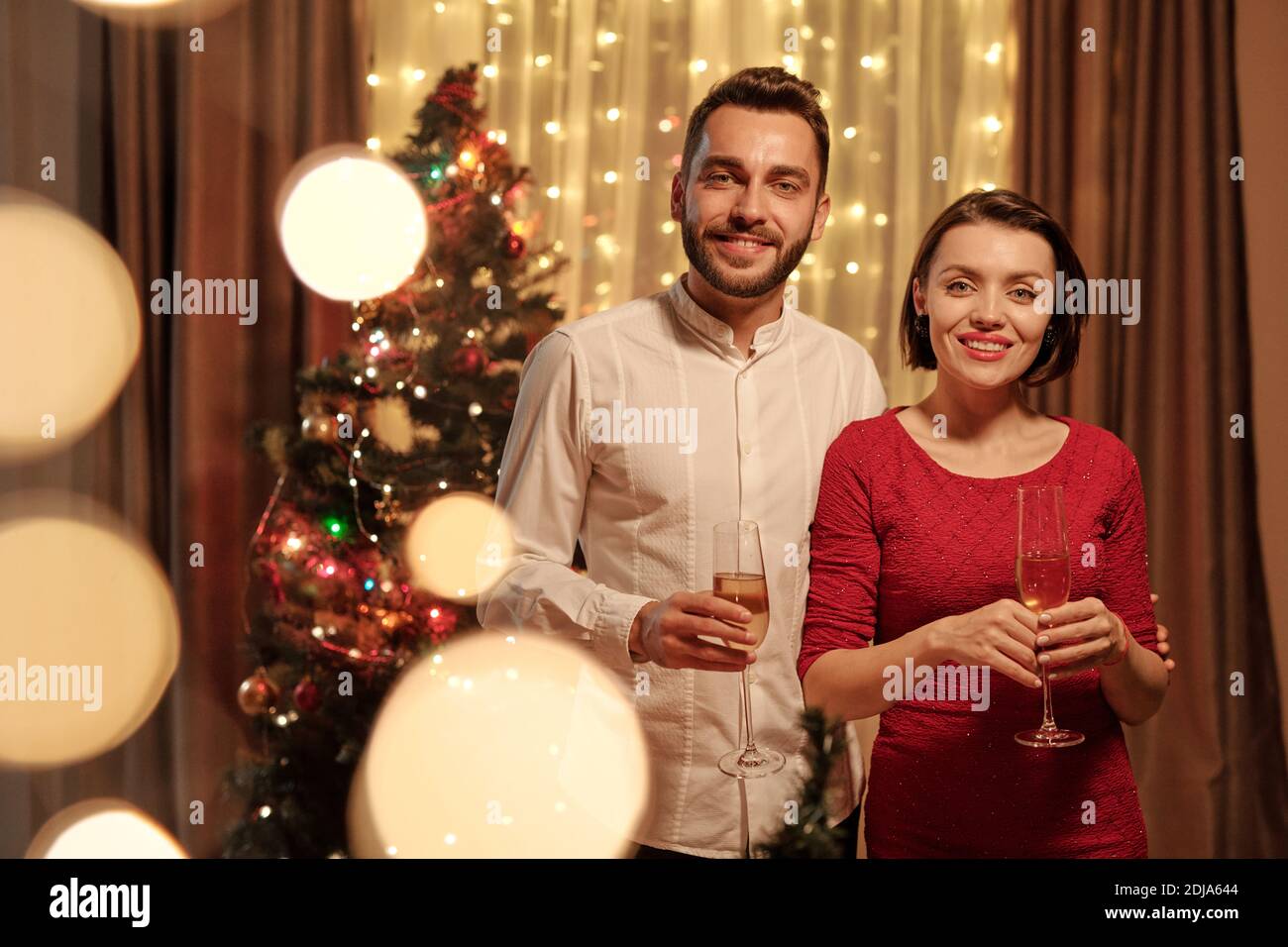 Portrait of smiling young couple in festive attires standing with champagne flutes against decorated tree, Christmas lights effect Stock Photo