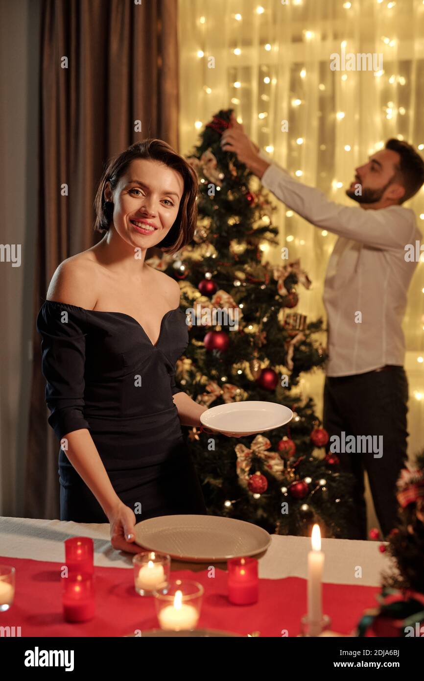 Portrait of happy attractive girl in fashionable dress putting plate on table while preparing for Christmas dinner with boyfriend Stock Photo