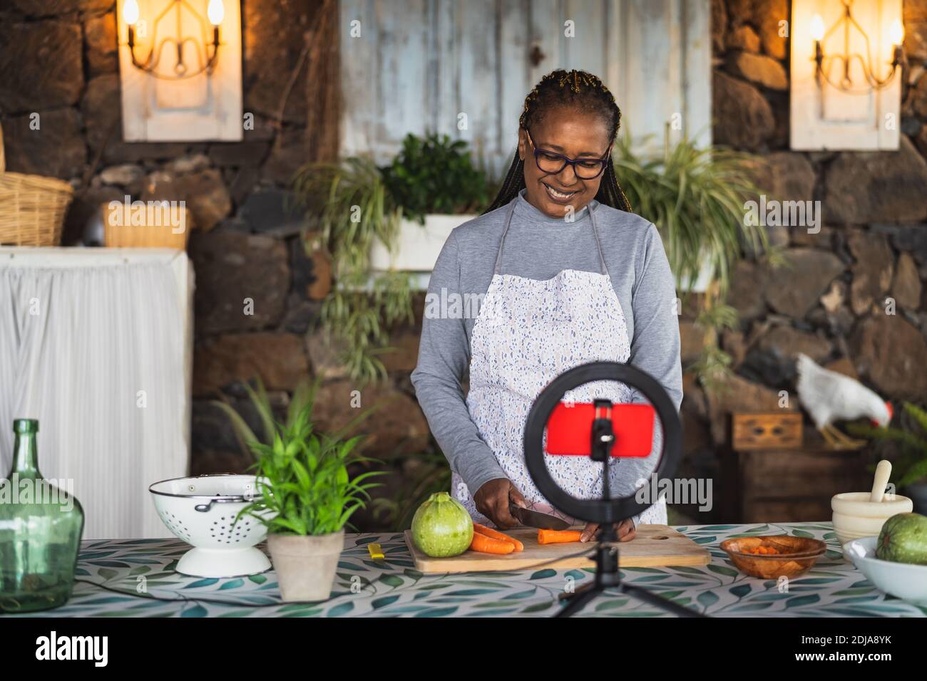 African senior woman preparing food recipe while streaming online with mobile smartphone cam for web kitchen masterclass channel Stock Photo