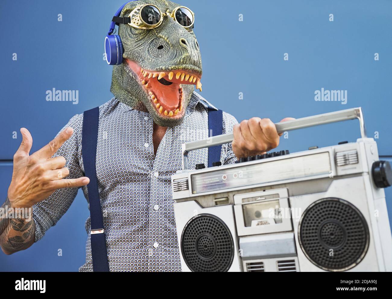 Crazy senior man having fun wearing t-rex mask while listening to music with headphones and vintage boombox stereo outdoor Stock Photo