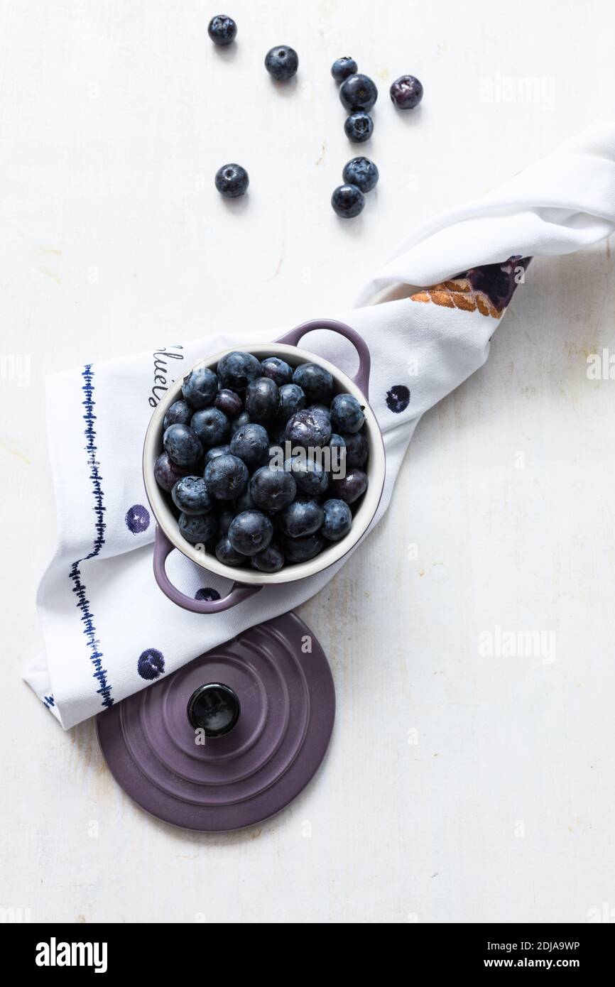 Elegant snack scene. A purple pot full of bluish delicious blueberries on a nice napkin. Some blueberries are isolated on the wooden table. Stock Photo
