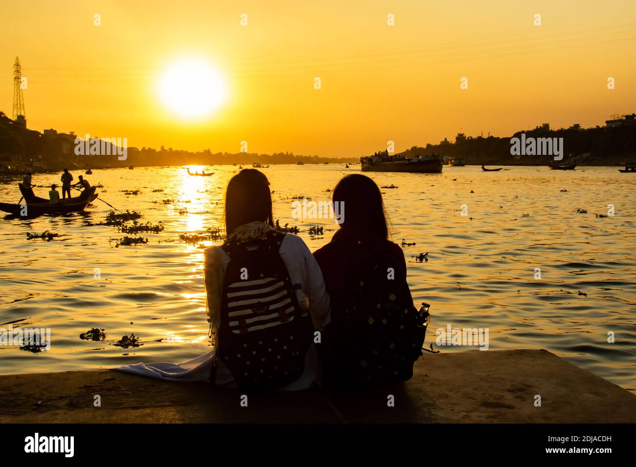 Silhouette of two students looking at the Buriganga river at the sunset, with tens boats crossing the river in Dhaka. Stock Photo
