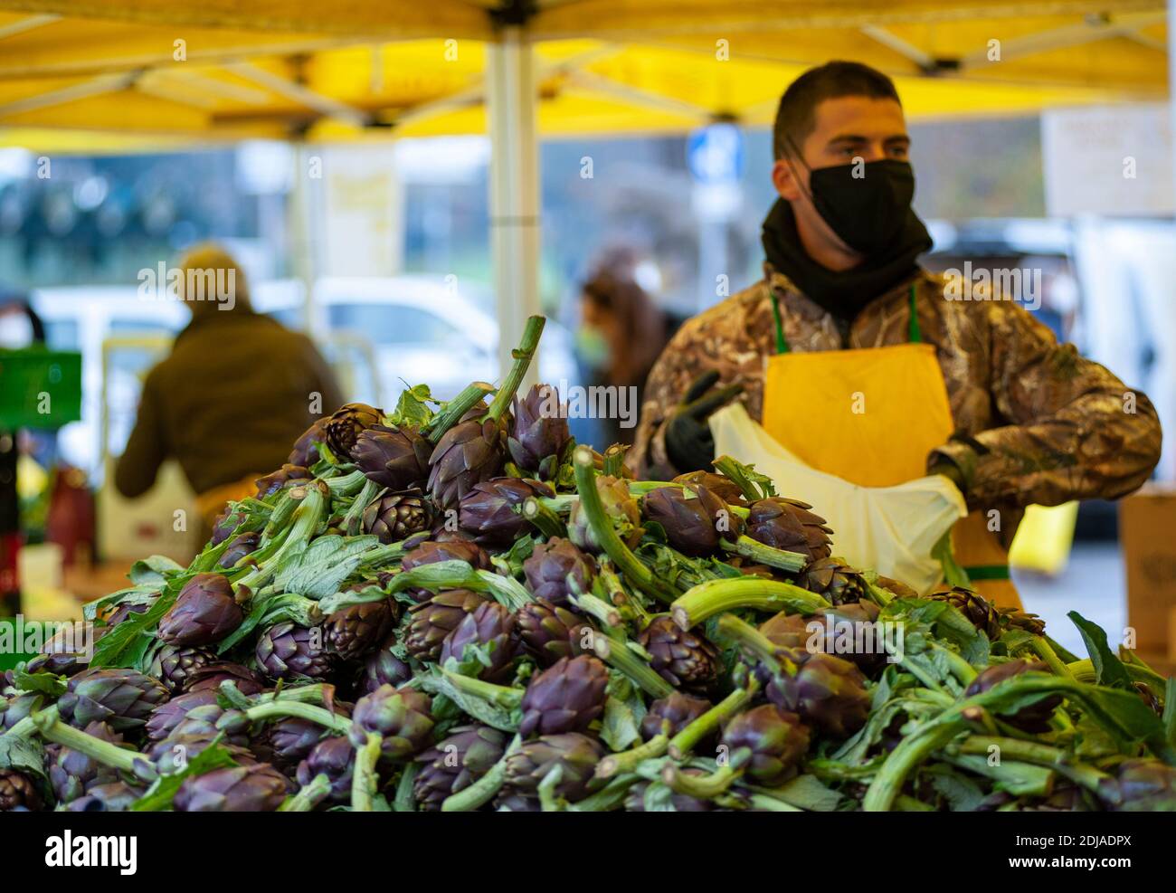 Greengrocer with black protective mask. A lot of artichokes in the foreground. Stock Photo