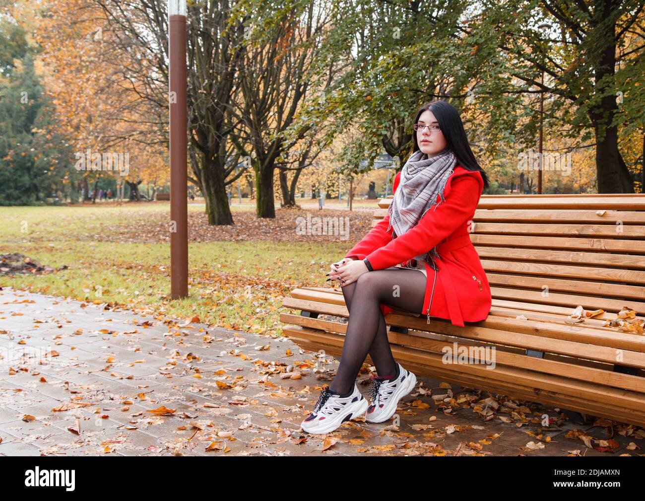 young beautiful girl in a red coat sitting on a bench in a city park ...