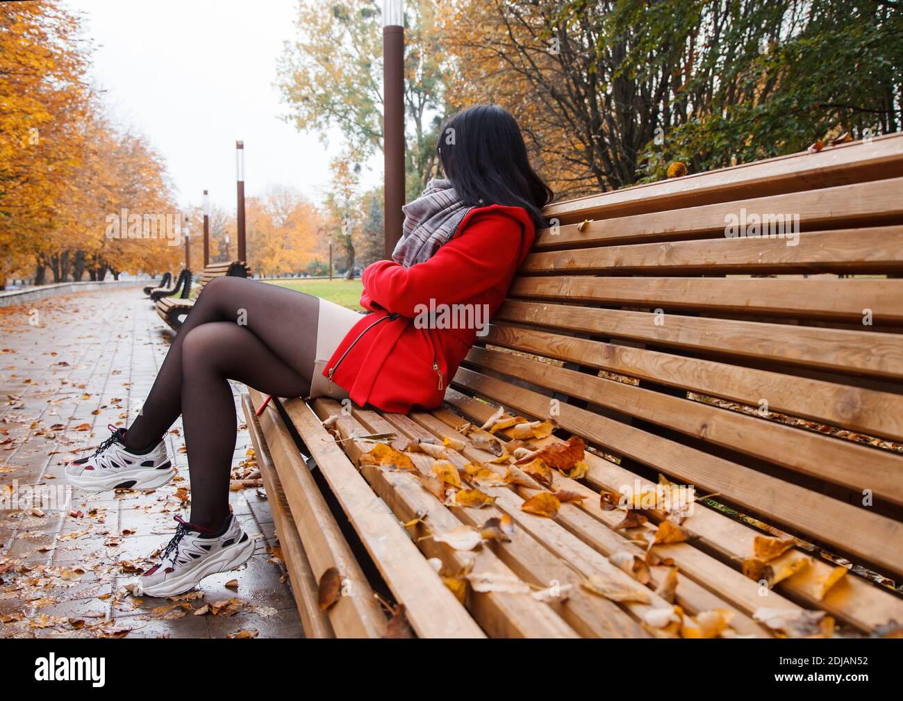 young beautiful girl in a red coat sitting on a bench in a city park ...
