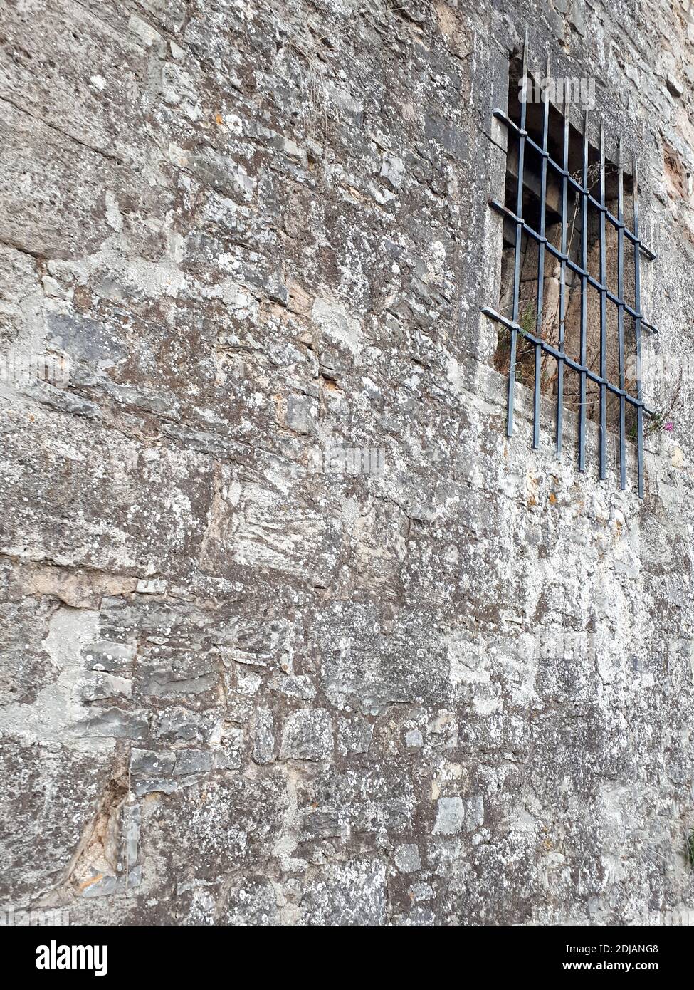 A vertical shot of an old stone building wall with a window with fences ...