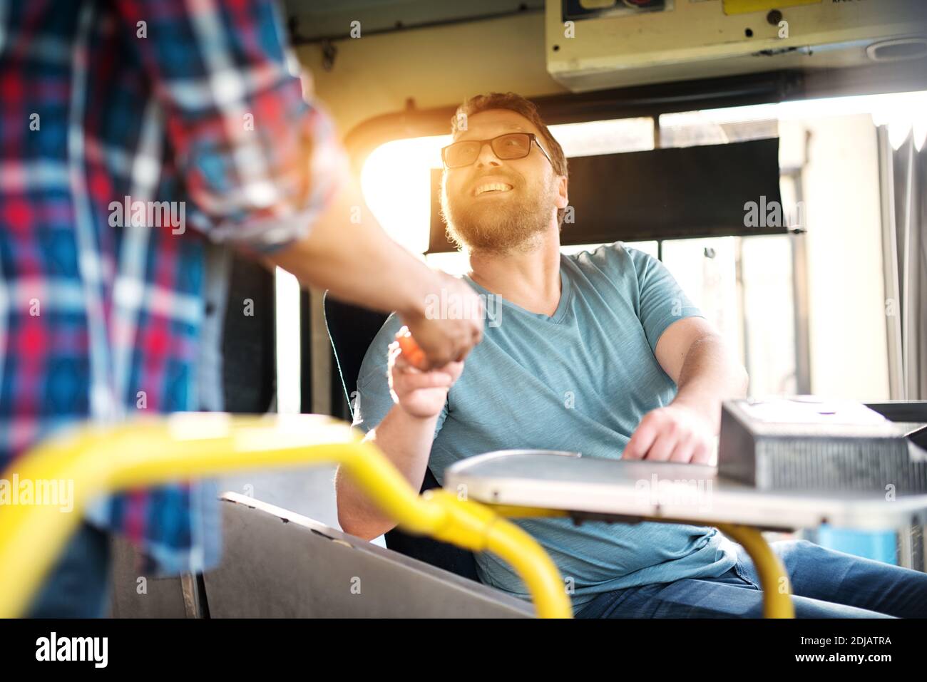 Cheerful young male bus driver is looking at the passenger and politely smiling while charging the ticket fee. Stock Photo