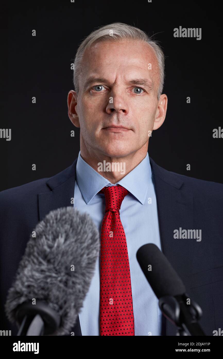 Vertical portrait of mature man standing at podium and speaking to microphone against black background Stock Photo