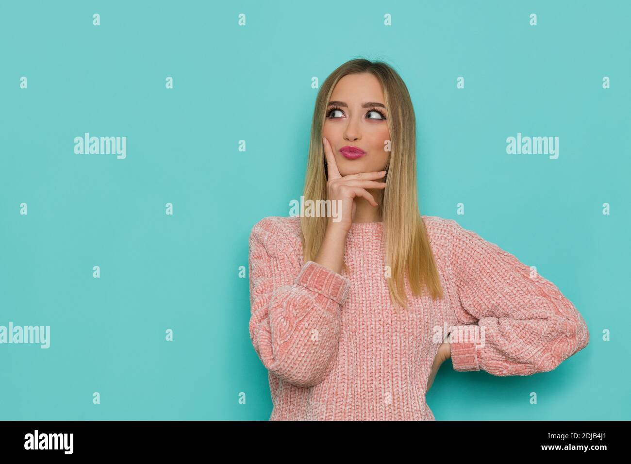 Excited young woman in pink sweater is holding hand on chin, grimacing and looking at the side. Waist up studio shot on turquoise background. Stock Photo