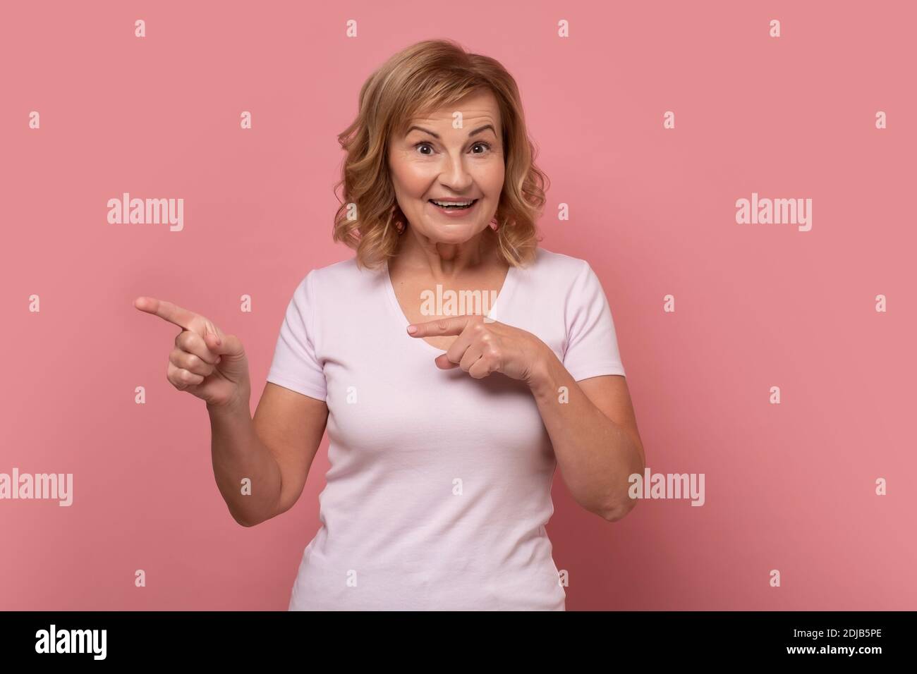 Check this out. Senior woman smiling cheerfully and pointing with forefingers away, indicating copy space on yellow wall, having joyful happy look Stock Photo