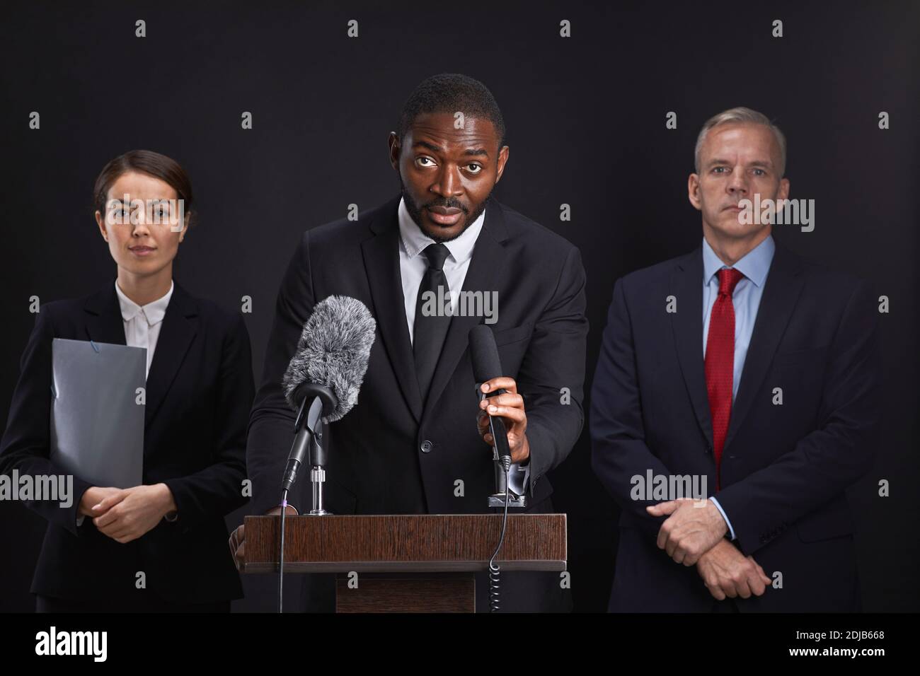 Waist up portrait of African-American man giving speech standing at podium with two assistants in background Stock Photo