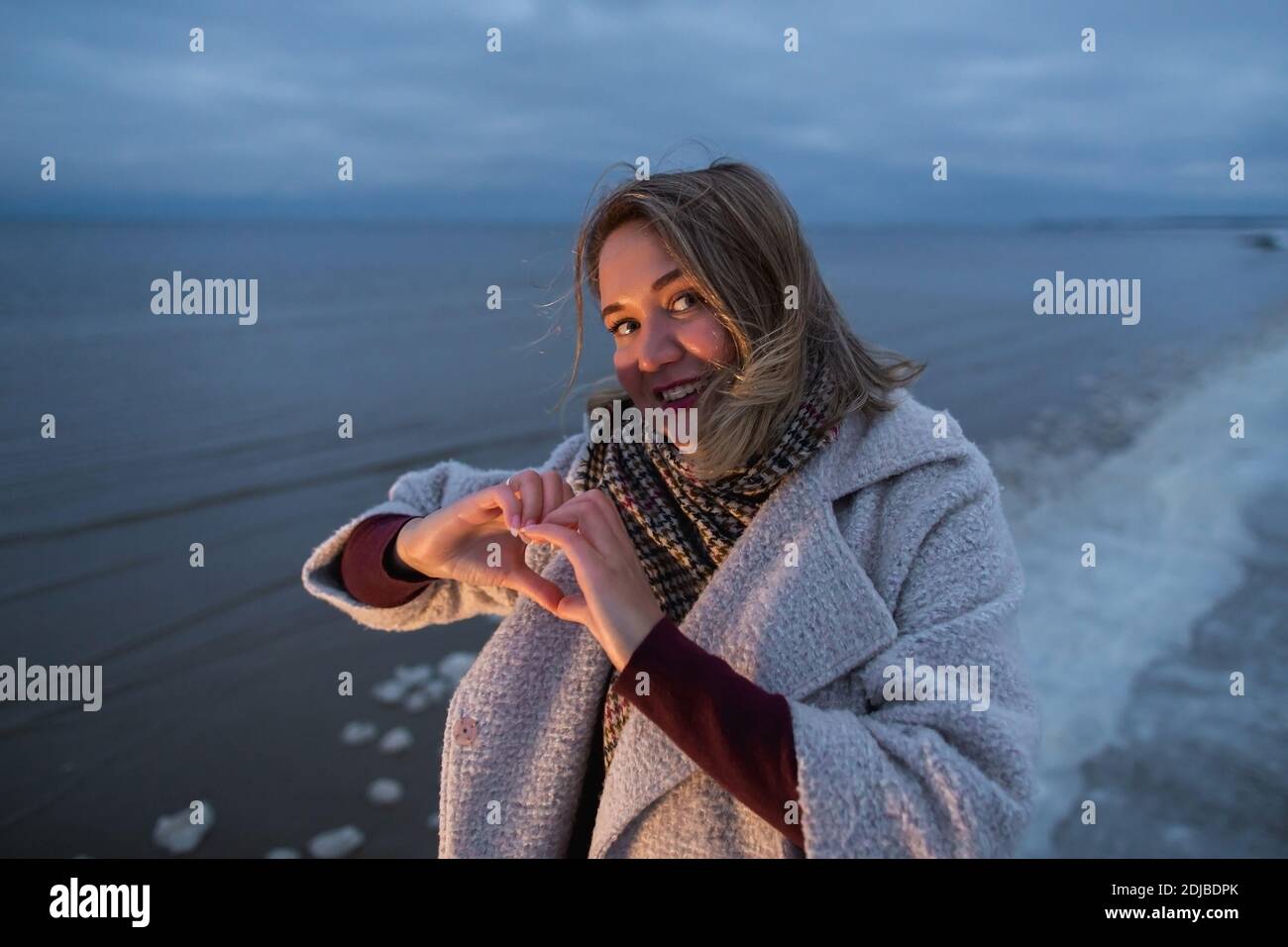 Beautiful woman showing heart shape of her hands on the winter sea coast evening time warm light. Stock Photo
