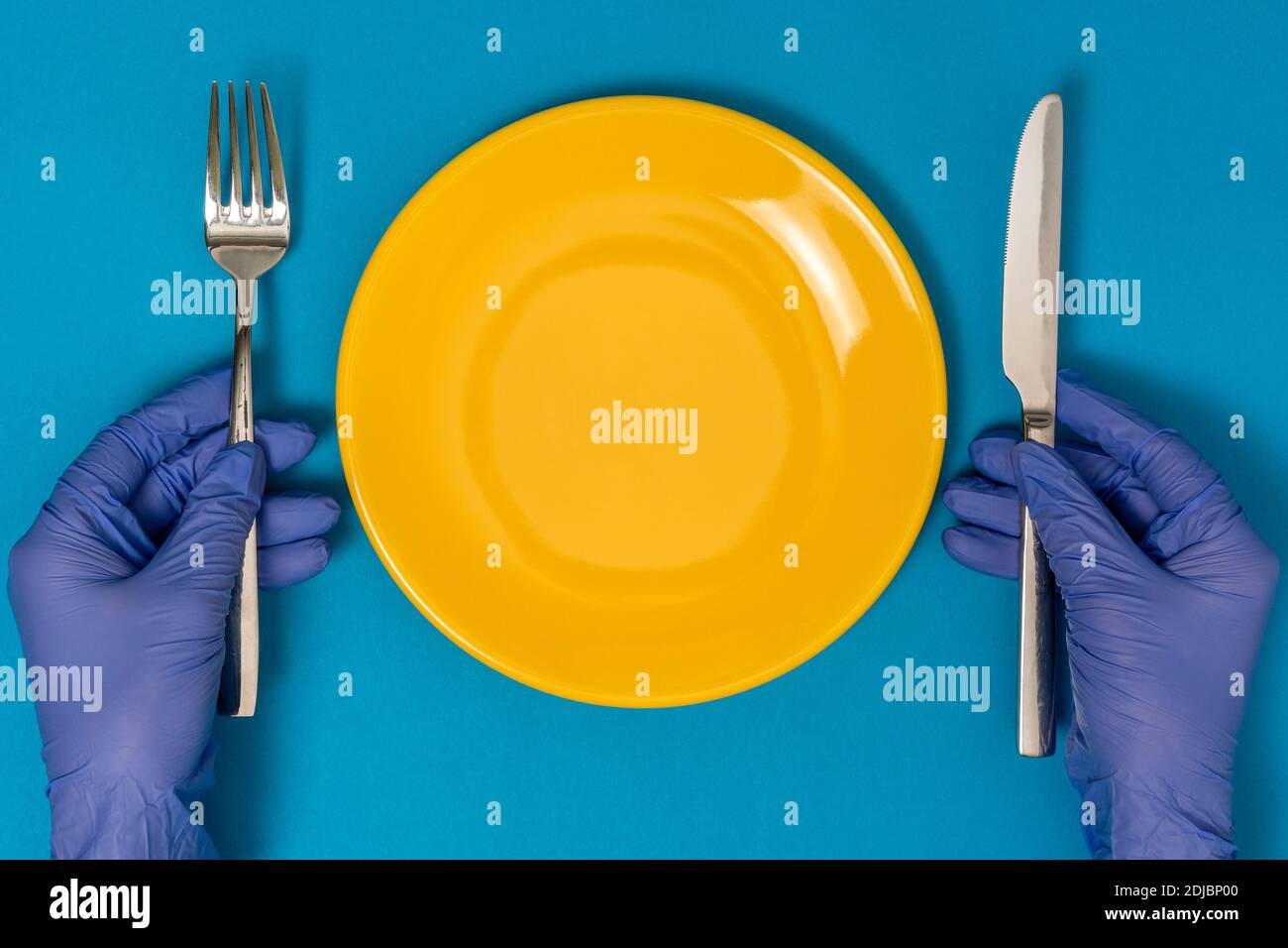 Hands in gloves keeping fork and knife over empty plate. Concept of the impact of the coronavirus pandemic on the food service industry. Stock Photo
