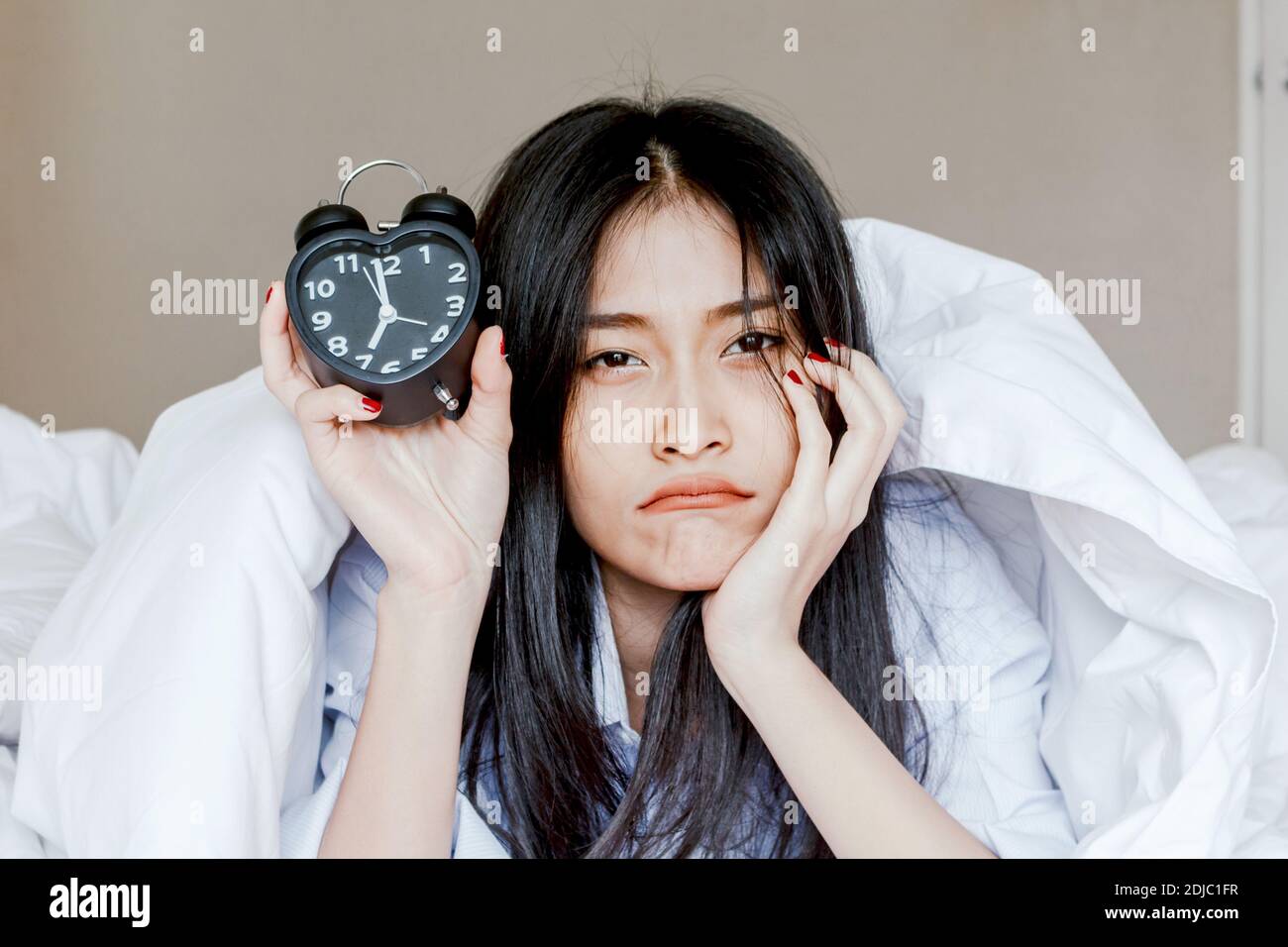 Portrait Of Sad Woman Showing Alarm Clock While Lying On Bed Stock