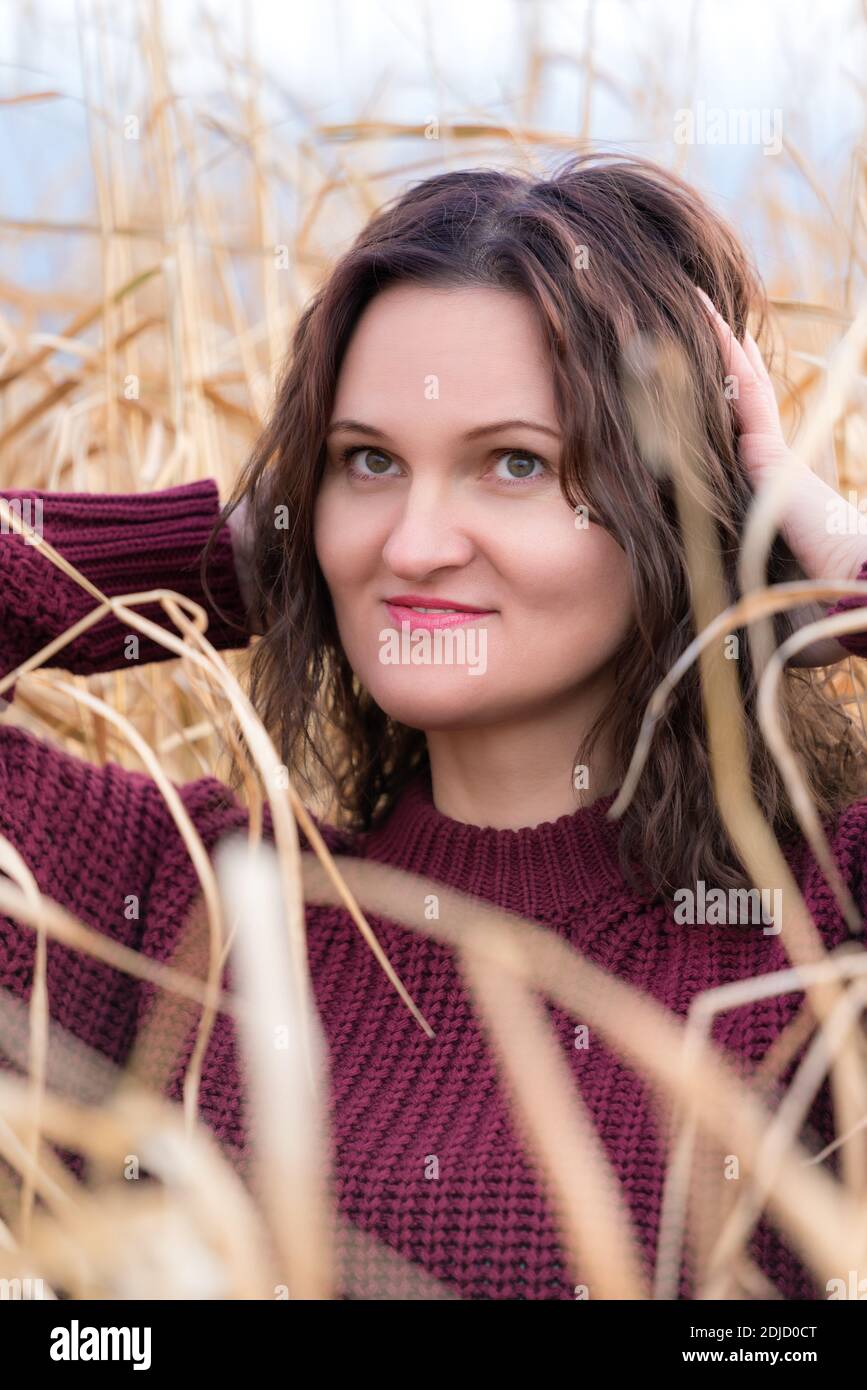Portrait of beauty young woman in brown sweater posing in dried grass in field. Cute woman with long curly brunette hair and red lips. Stock Photo