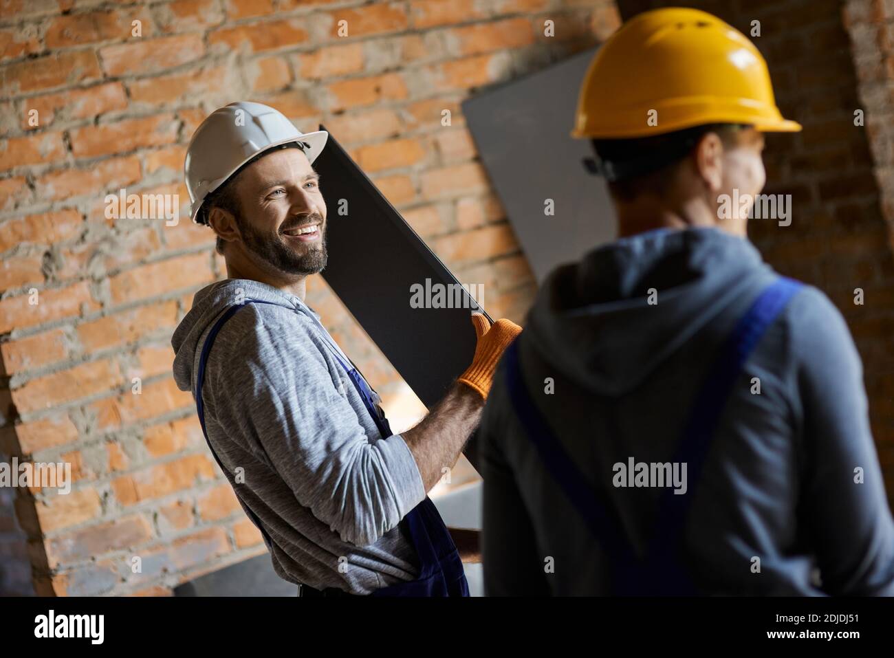 Cheerful young male builders wearing hard hats smiling, holding metal stud for drywall while working together at construction site. Building house, profession, teamwork concept Stock Photo