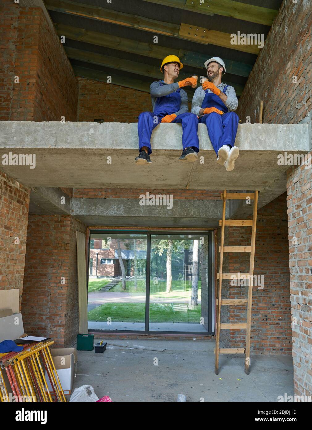 Two happy young builders in blue overalls and hard hats resting, sitting on the concrete floor, drinking coffee while working on house construction. Building house, partnership concept Stock Photo