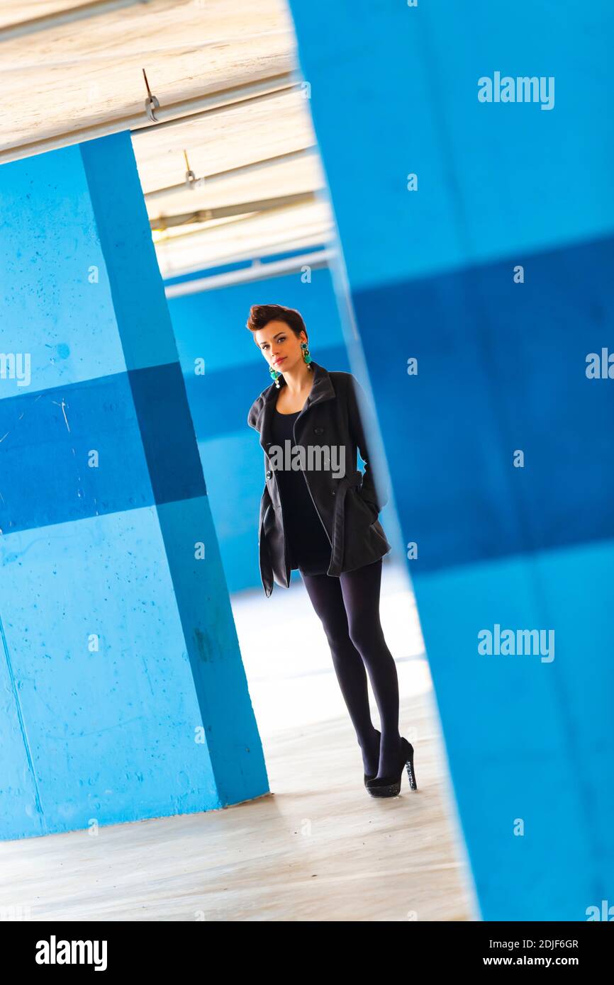 Teengirl in empty parking lot with Blue walls legs heels eyeshot eyes eye contact looking at camera tight space serious unsmiling Stock Photo