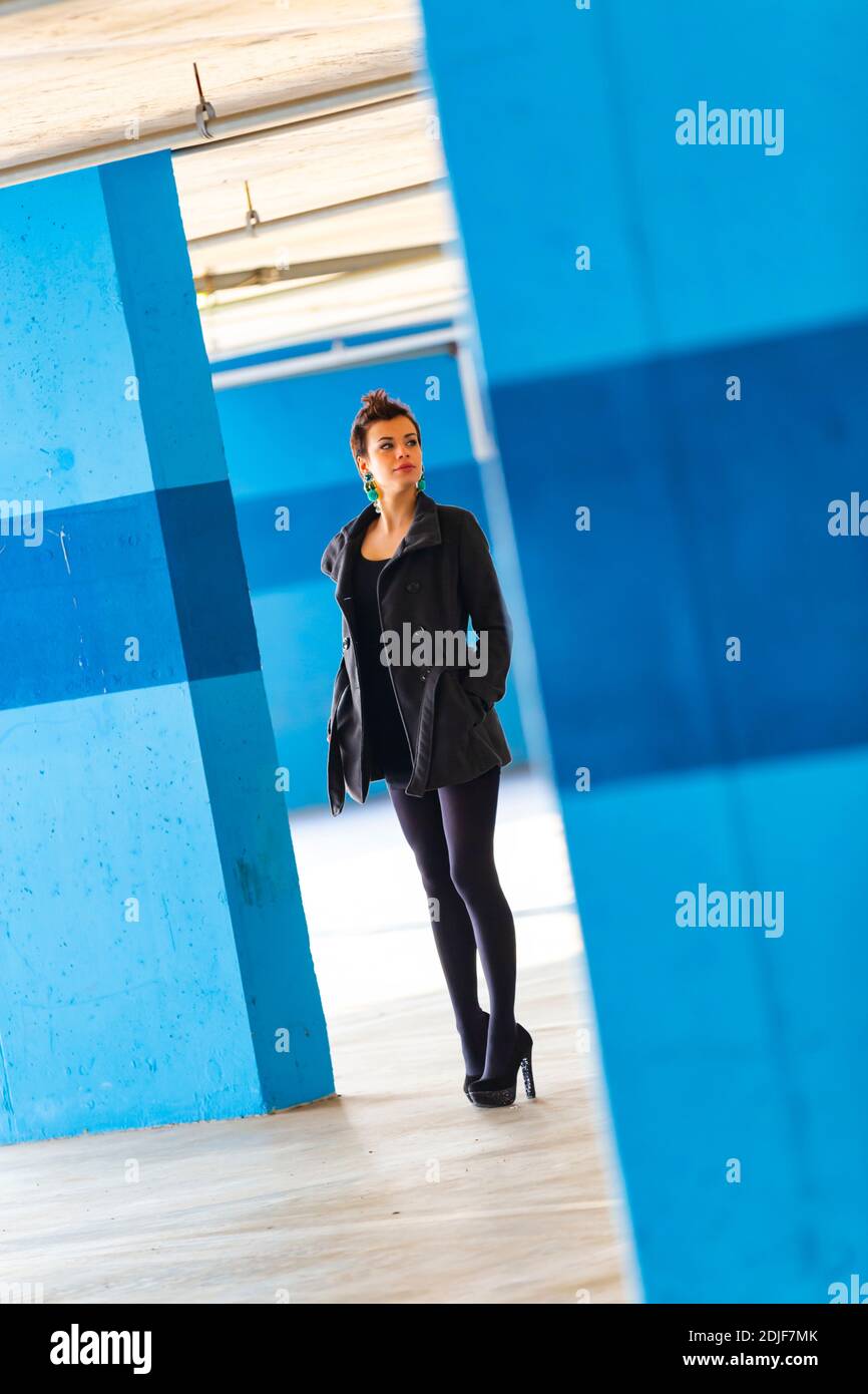 Teengirl in empty parking lot with Blue walls legs heels tight space serious unsmiling looking away aside back Stock Photo