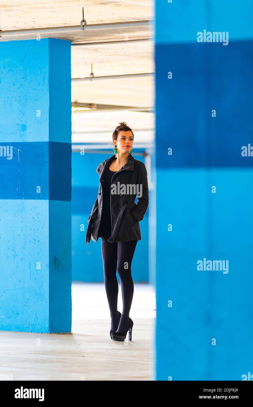 Teengirl in empty parking lot with Blue walls legs heels tight space serious unsmiling looking away aside back Stock Photo