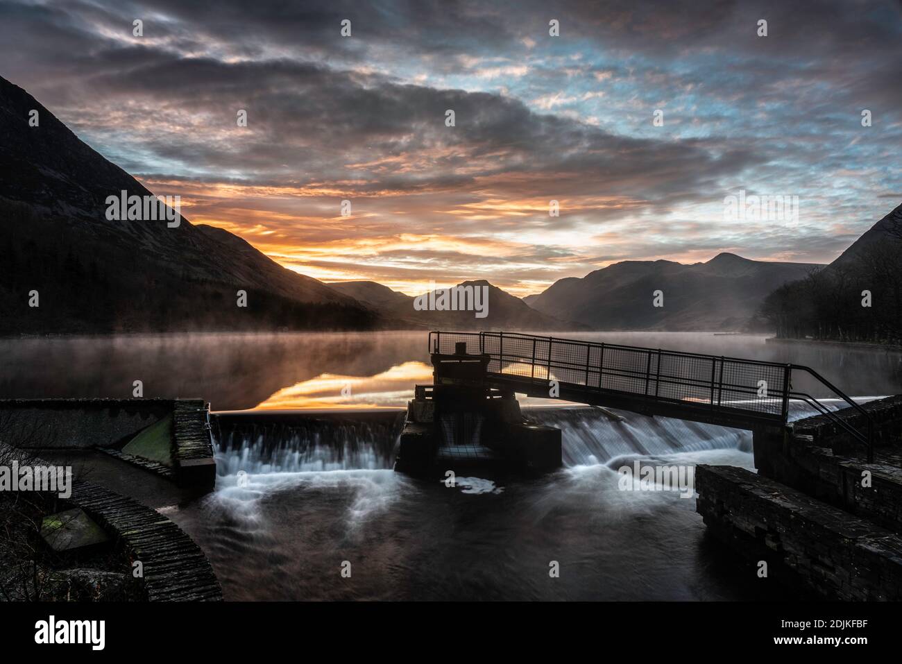 A beautiful winter sunrise at the weir at Crummock Water, with mist and a golden glow on clouds reflected in the lake. Lake District, Cumbria, England Stock Photo